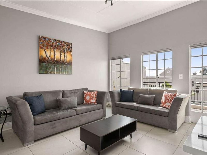 A bright living room featuring neutral tiled flooring, crown molding, and multiple grid-pattern windows providing natural light and access to an outdoor area.