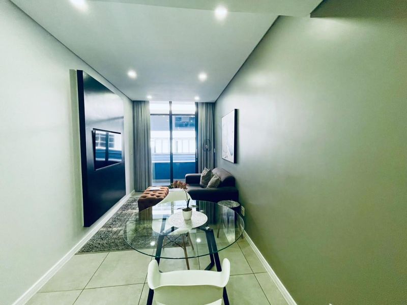 Modern open-plan living area featuring tiled floors, recessed lighting, and a feature wall, with direct access to a balcony through sliding glass doors.