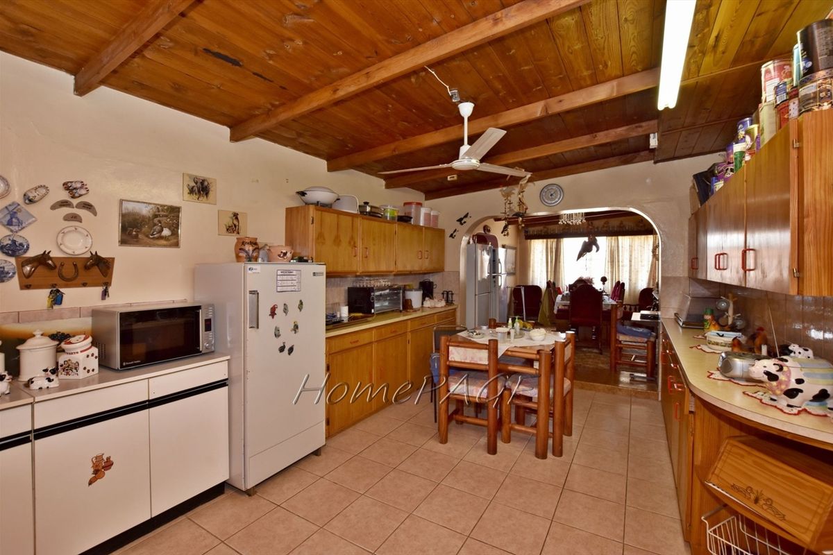 A dated kitchen featuring wooden cabinetry, tiled flooring, and exposed timber ceiling beams, with an open-plan layout leading into the dining area.