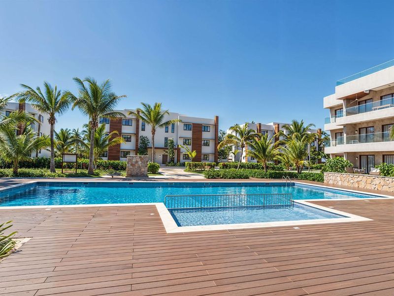 A well-maintained communal swimming pool area featuring a large pool, a smaller wading pool, and extensive timber decking surrounded by landscaped gardens and apartment buildings.
