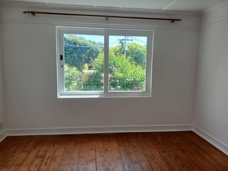 A bright, empty bedroom featuring polished timber flooring, a sliding window with garden views, and classic picture rail detailing.
