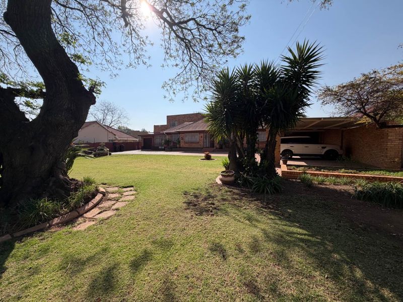 A spacious front exterior view featuring a large lawn, mature trees, a paved driveway, and a covered carport area.