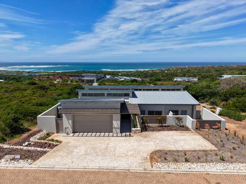 Aerial view of a modern coastal home featuring a metal roof, paved driveway, double garage, and stone accents, set against a scenic ocean backdrop.