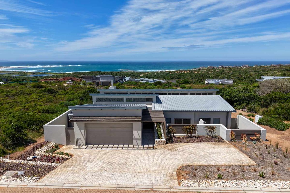 Aerial view of a modern coastal home featuring a metal roof, paved driveway, double garage, and stone accents, set against a scenic ocean backdrop.