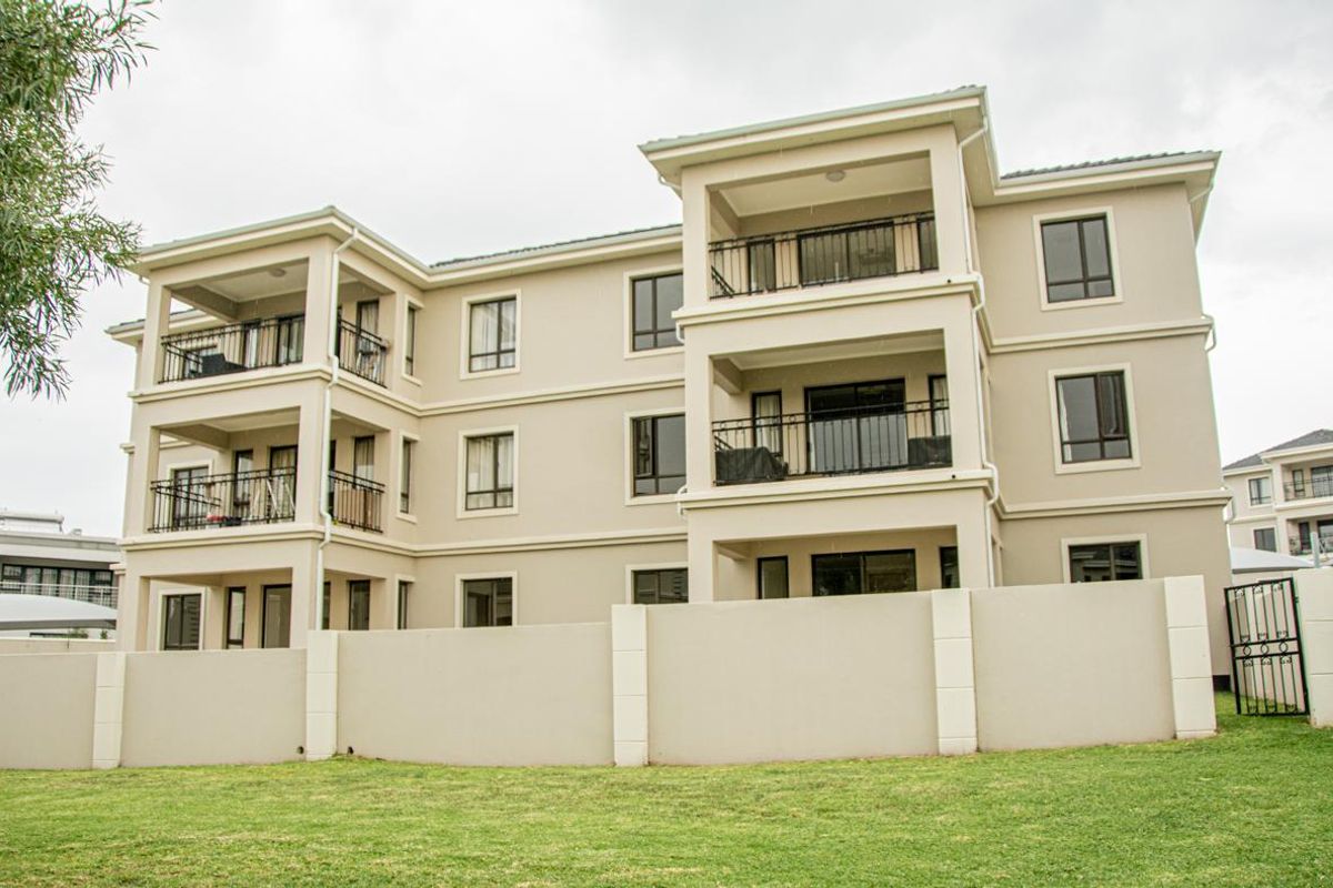 Rear view of a multi-story apartment complex featuring private balconies with wrought iron railings, a perimeter wall, and a landscaped lawn area.