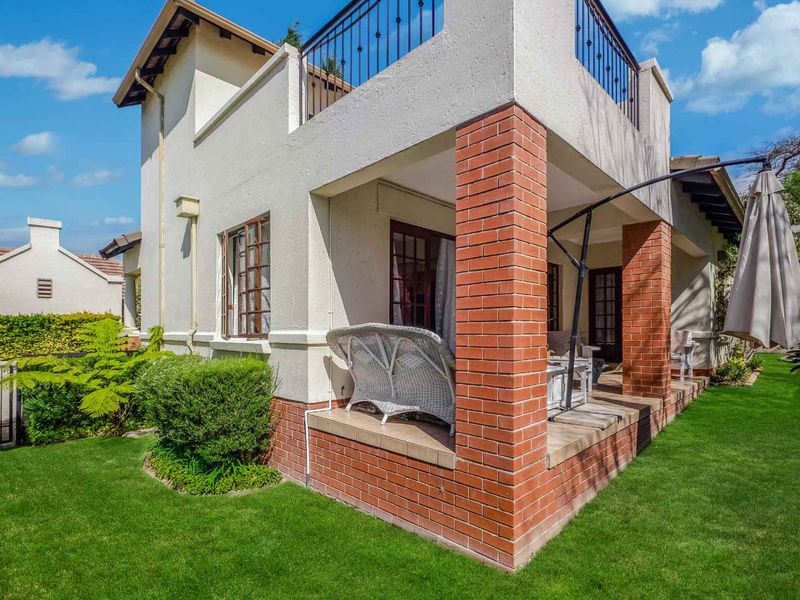 Rear view of the townhouse featuring a covered patio with brick pillars, a second-story balcony with metal railings, and a well-maintained garden.