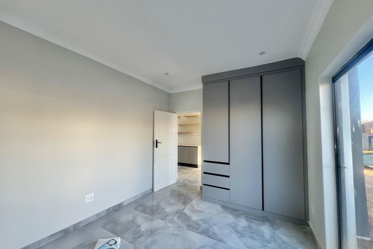 A modern bedroom featuring sleek grey built-in cupboards, large format floor tiles, and recessed ceiling lighting, with a view into the adjacent kitchen area.