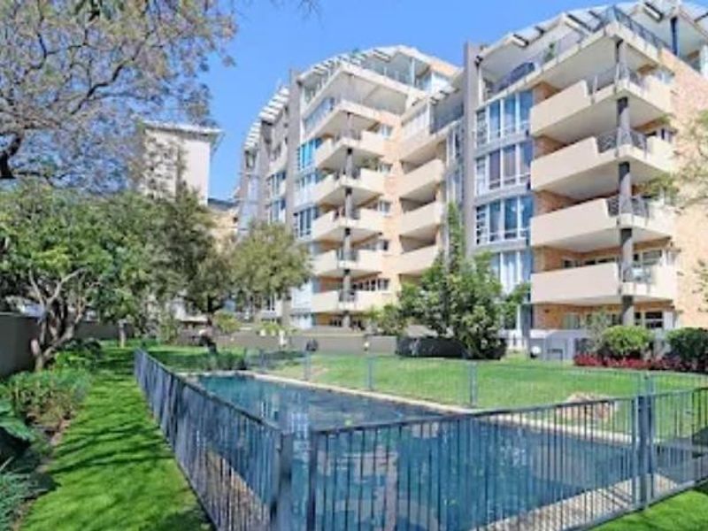 Rear view of the apartment complex featuring a swimming pool, secure pool fencing, landscaped garden areas, and private balconies for each unit.