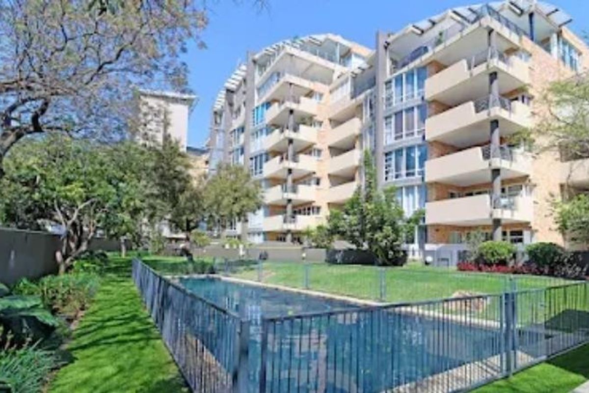 Rear view of the apartment complex featuring a swimming pool, secure pool fencing, landscaped garden areas, and private balconies for each unit.