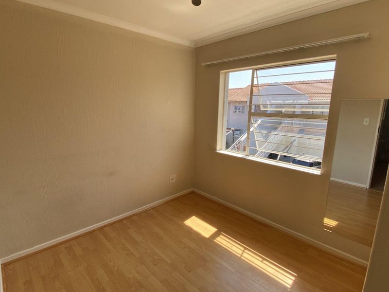 An empty bedroom featuring laminate flooring, neutral walls, and a large window with louvre panes, offering a blank canvas for personalization.
