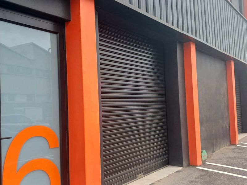 Modern industrial unit exterior featuring a secure roller shutter door, glass entrance, and dedicated parking bay with contemporary orange and dark grey facade.