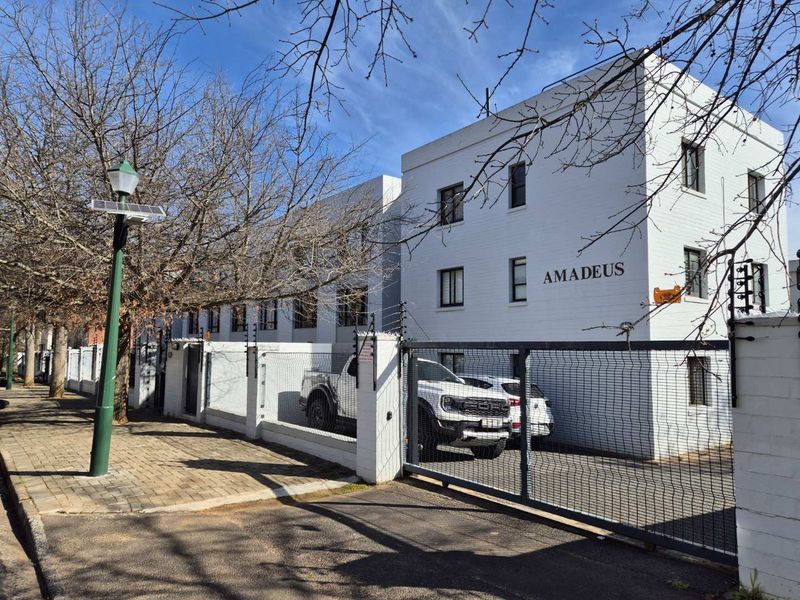 Front exterior view of the Amadeus apartment complex, featuring a secure gated entrance, white painted brick facade, and perimeter electric fencing.