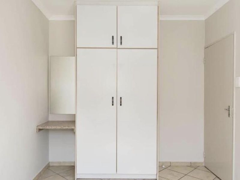 A clean bedroom interior featuring a white built-in wardrobe with overhead storage and an integrated vanity shelf, finished with neutral tiled flooring.