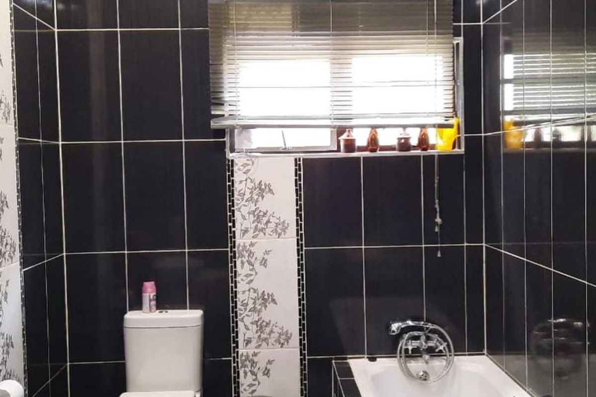 A compact bathroom featuring dark wall and floor tiles, a built-in bathtub, a standard toilet, and a window with fitted blinds.