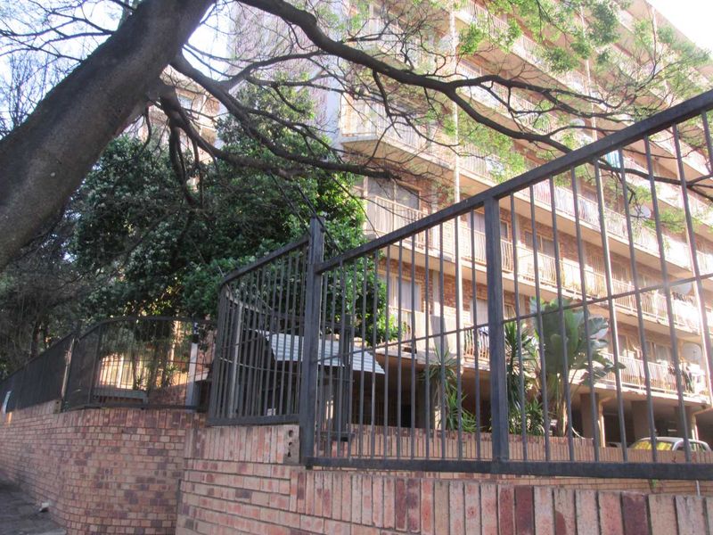 Exterior view of a multi-story apartment building featuring brick boundary walls, metal security fencing, and private balconies.