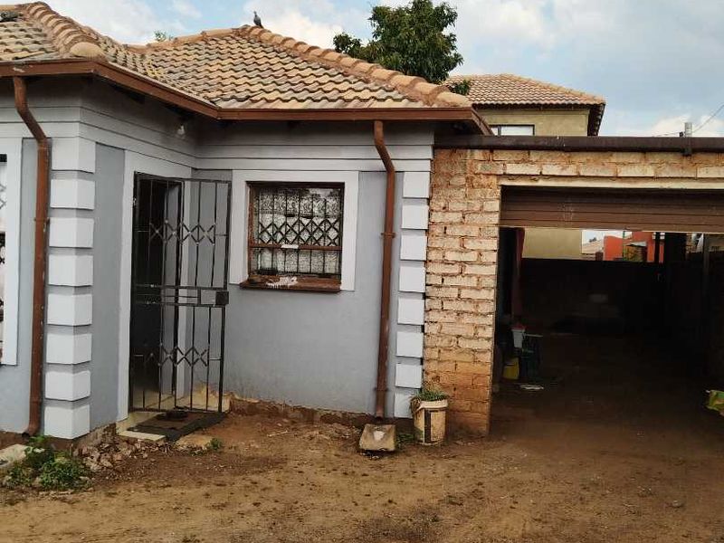Front view of a single-story house featuring a tiled roof, security gates, and an attached brick garage with an unpaved driveway area.