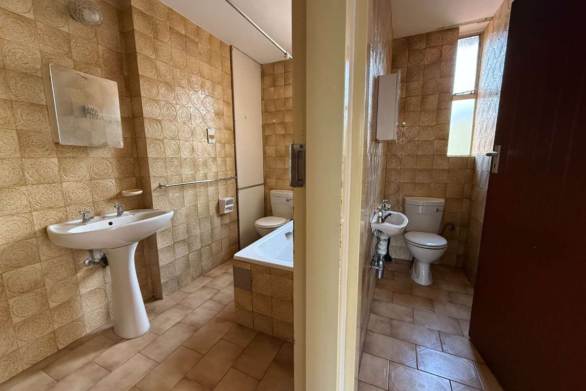 A dated bathroom featuring a pedestal basin, built-in bathtub, and separate toilet area with original ceramic tiling throughout.