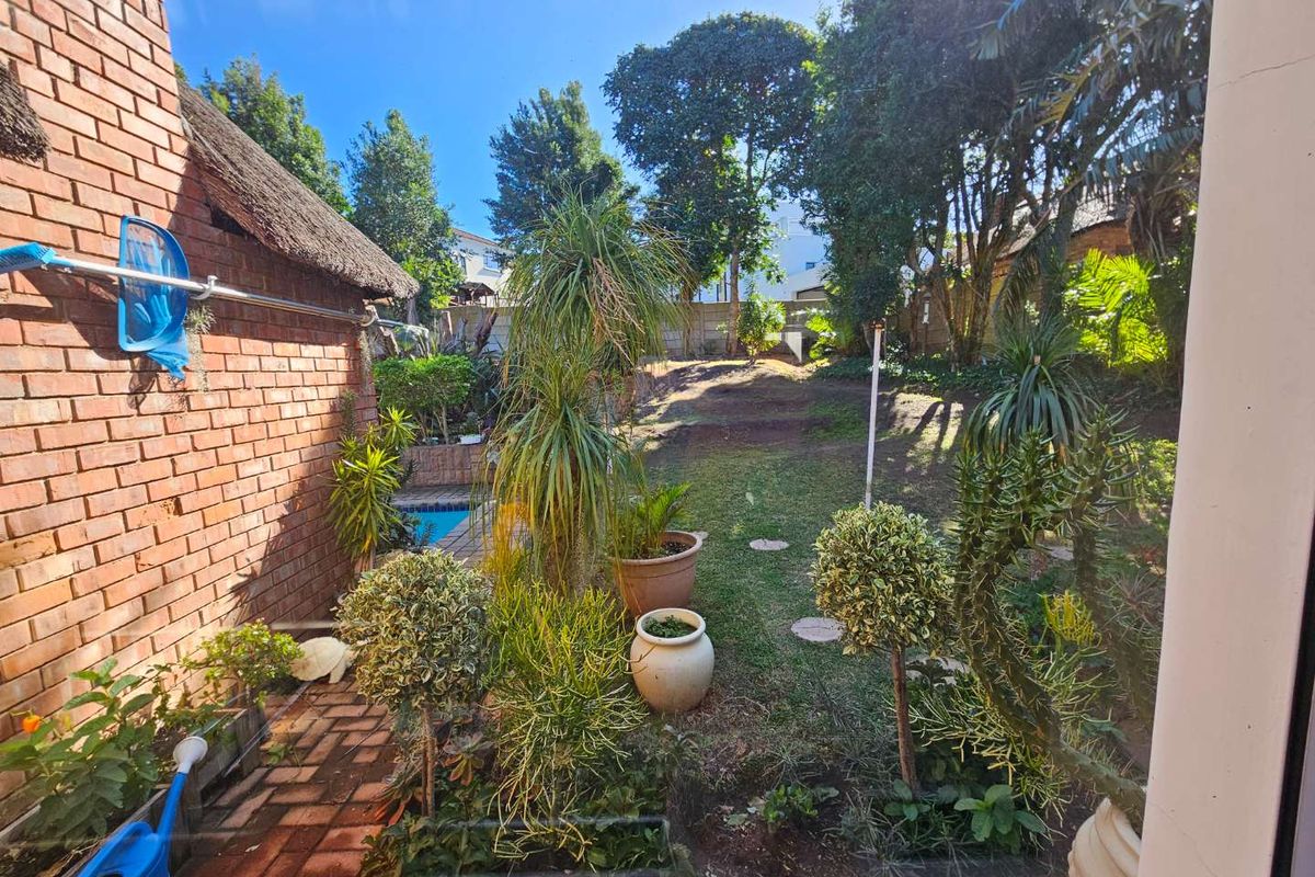 A rear garden view featuring a brick exterior wall, thatched roof section, paved patio area, and a swimming pool set within a landscaped backyard.