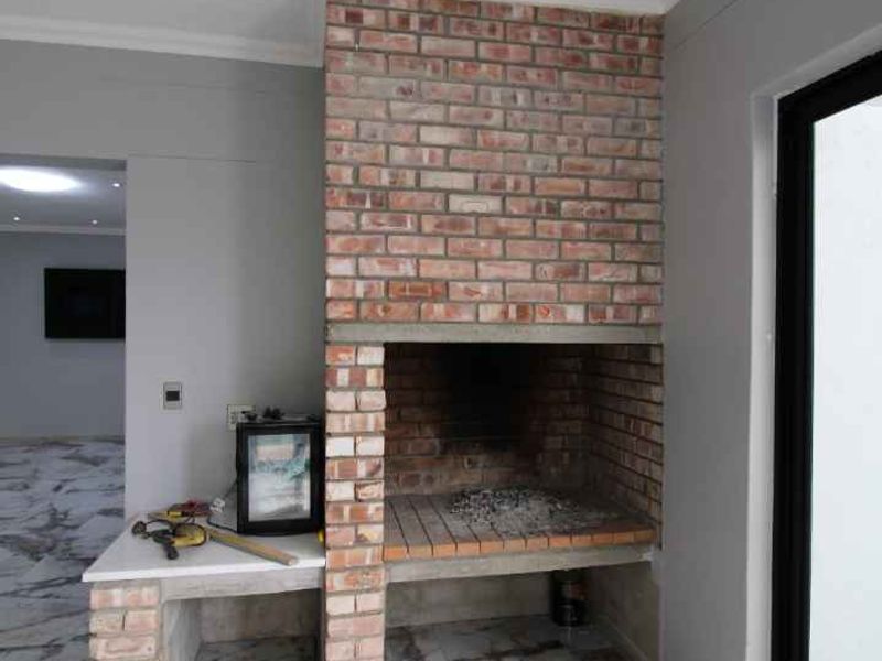 A modern covered patio area featuring a built-in brick braai, marble-look tiled flooring, and large sliding glass doors leading to the interior.