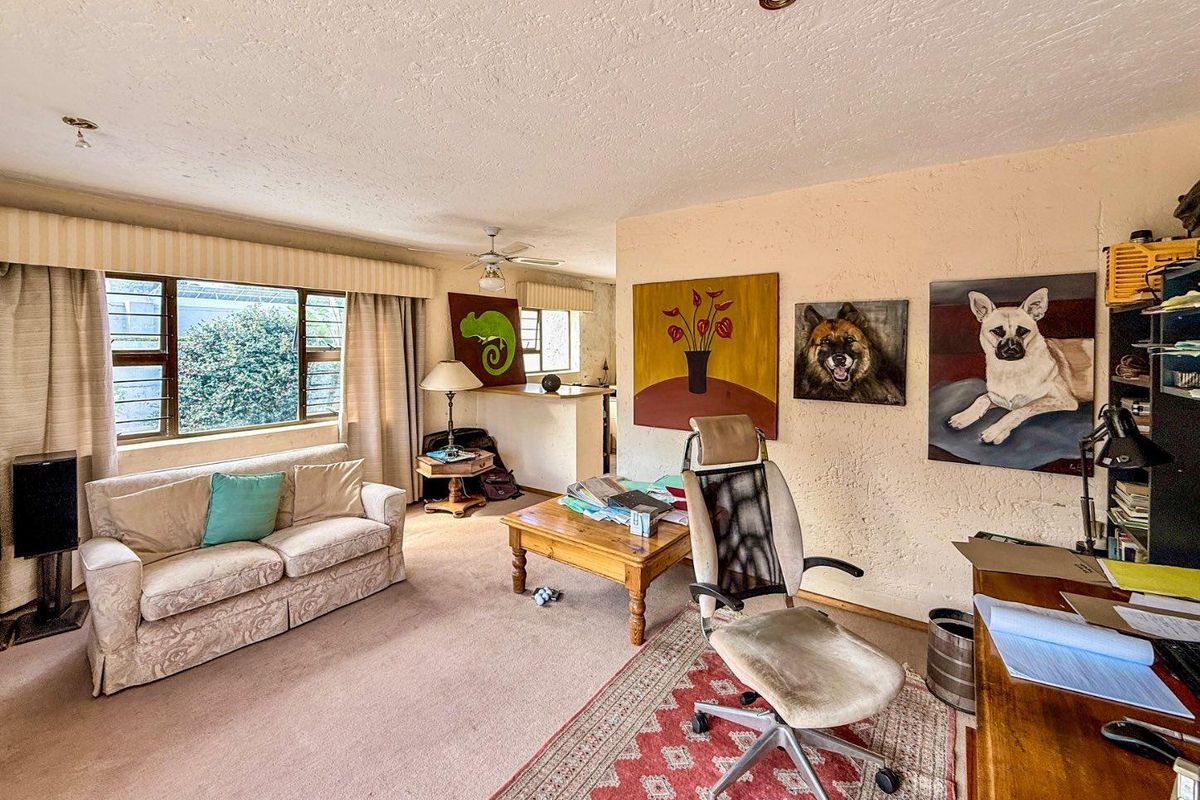 A multi-functional living area featuring carpeted flooring, large windows, and a semi-open plan layout connecting to an adjacent space.