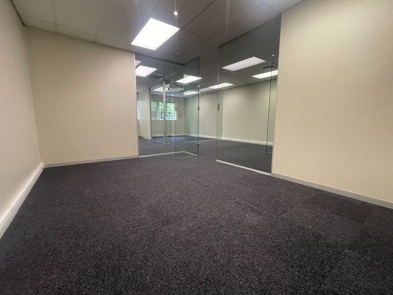 A clean, empty office space featuring modern glass partitions, recessed ceiling lighting, and dark grey carpeting, offering a professional and versatile work environment.