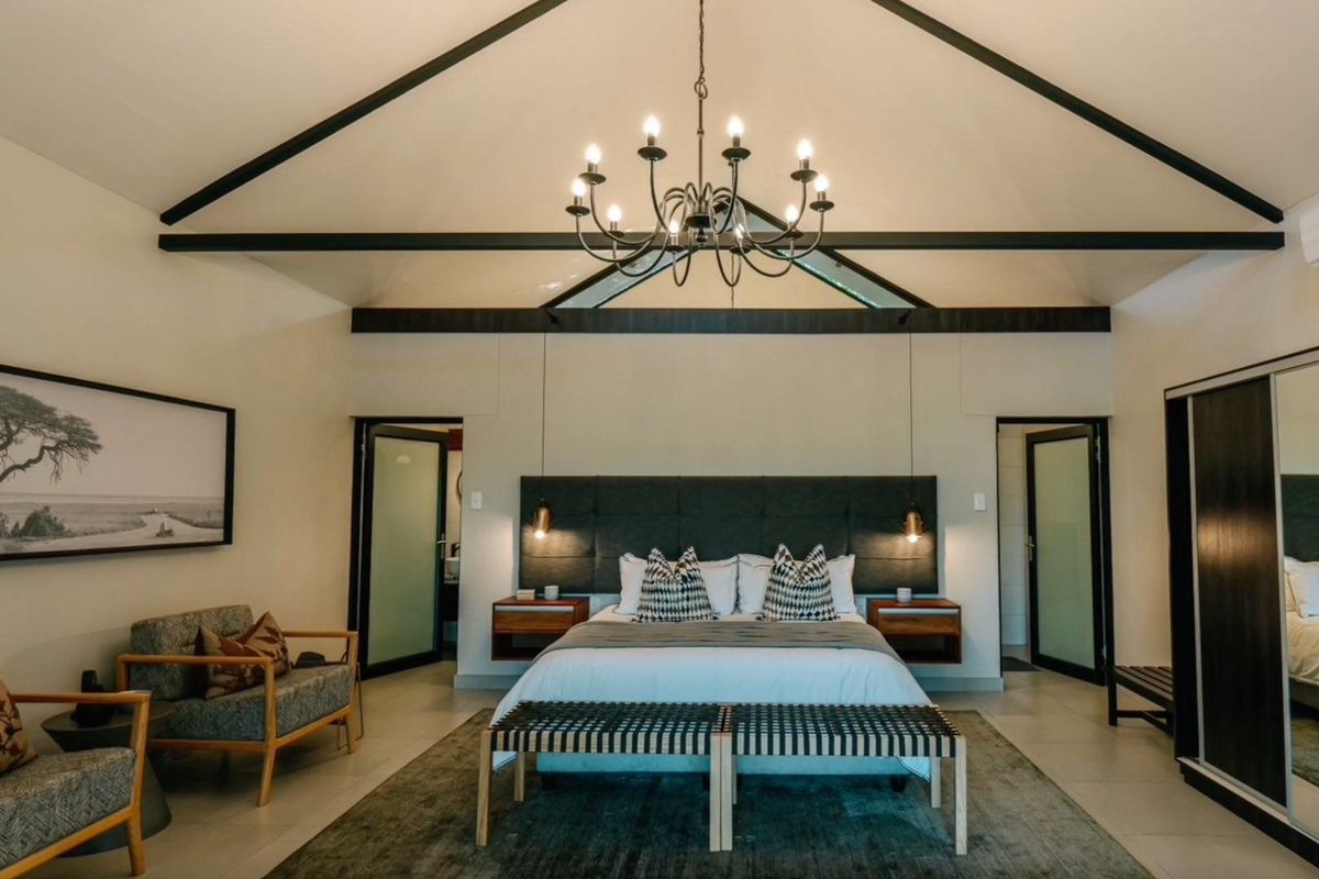 Spacious bedroom featuring a vaulted ceiling with exposed beams, a central chandelier, and integrated bedside lighting, offering a modern and comfortable lodge atmosphere.