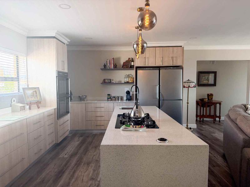 Modern open-plan kitchen featuring a central island with stone countertops, built-in oven, gas cooktop, and contemporary light wood cabinetry with floating shelves.
