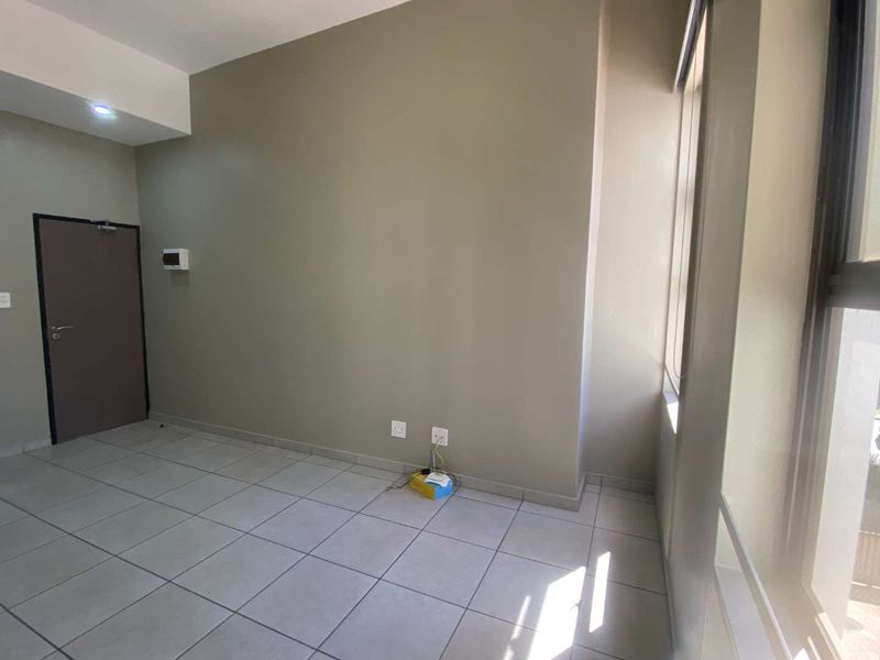 A bright, empty living area featuring neutral wall tones, tiled flooring, and large windows that provide ample natural light.