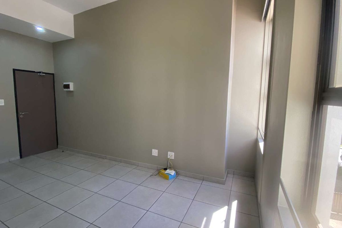 A bright, empty living area featuring neutral wall tones, tiled flooring, and large windows that provide ample natural light.