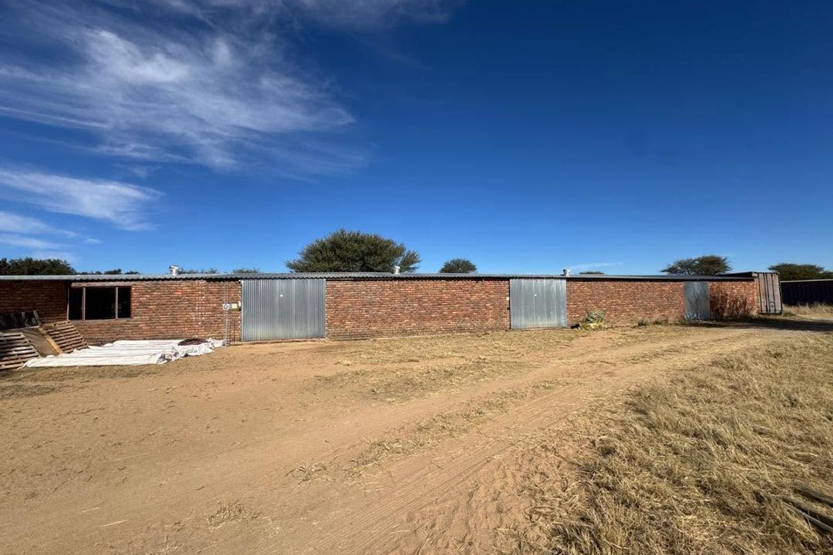 A long, single-story brick outbuilding or storage structure featuring multiple metal roller doors and a corrugated roof, situated on a large, unpaved plot.