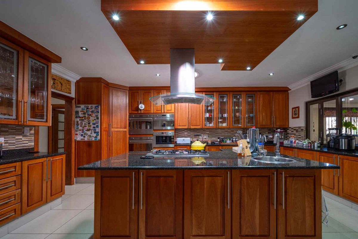Spacious kitchen featuring warm wooden cabinetry, a large central island with granite countertops, built-in appliances, and a modern stainless steel extractor fan.