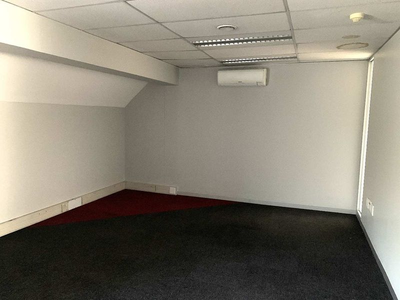 An empty office space featuring a suspended ceiling, wall-mounted air conditioning unit, and dark carpeting.