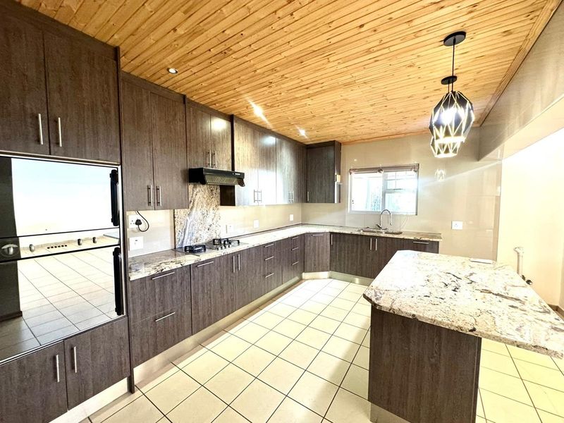 Modern kitchen featuring dark wood cabinetry, granite countertops, a central island, built-in oven, gas hob, and a distinctive timber-clad ceiling.