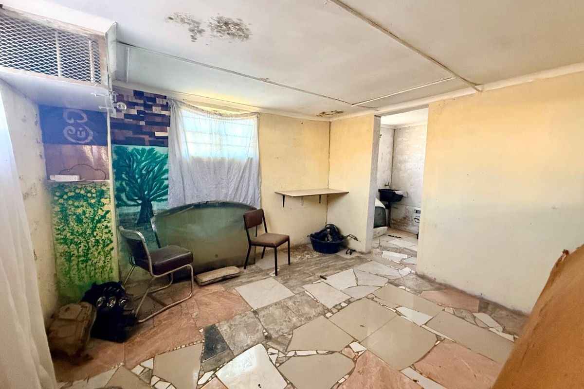 A dated interior space featuring irregular floor tiling, wall-mounted shelving, and a connected utility area, requiring significant renovation and repair.