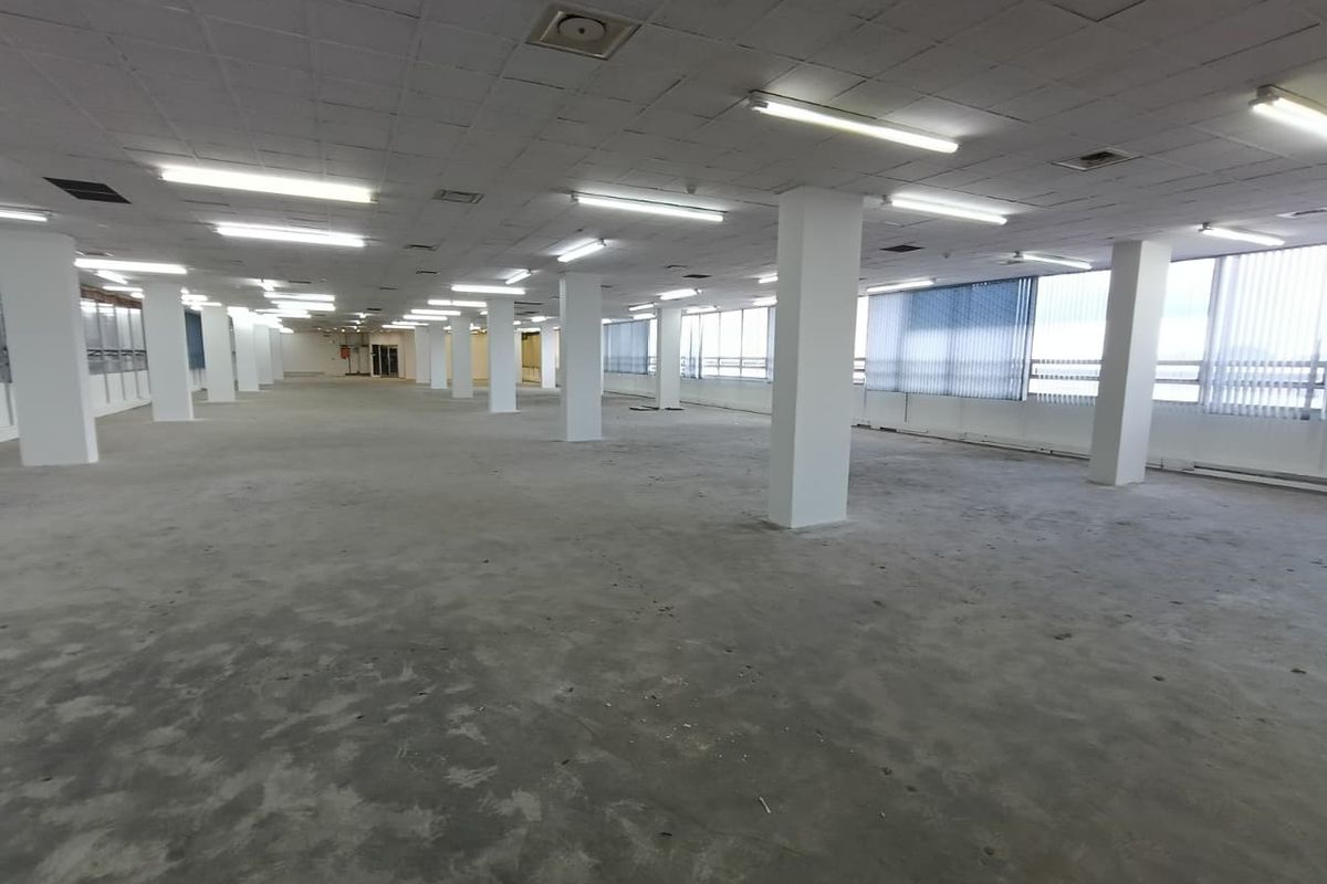 A large, open-plan office space featuring structural columns, suspended ceilings with fluorescent lighting, and expansive windows with vertical blinds.
