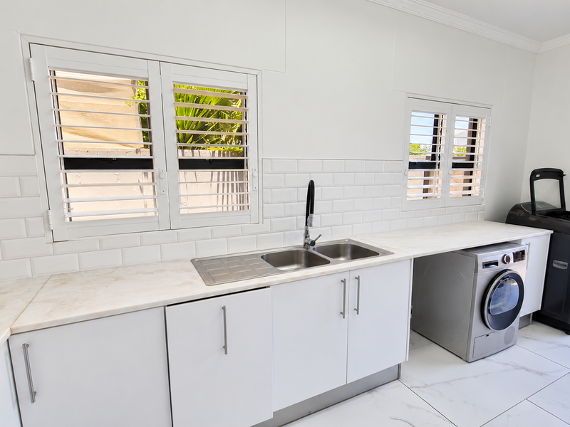 A clean, modern laundry room featuring white cabinetry, subway tile backsplash, marble-look countertops, a stainless steel double sink, and plantation shutters.