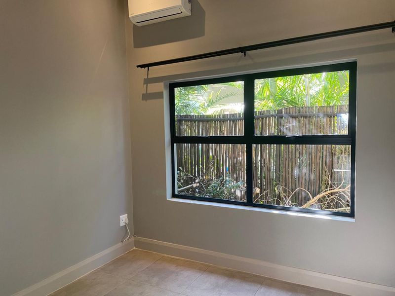 A clean, empty bedroom featuring neutral grey walls, tiled flooring, a modern black-framed window, and a wall-mounted air conditioning unit.