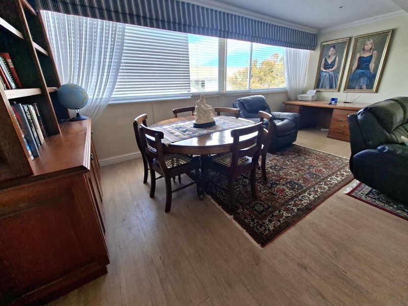 A spacious living area featuring light-toned laminate flooring, a built-in desk workspace, and large windows with integrated venetian blinds.