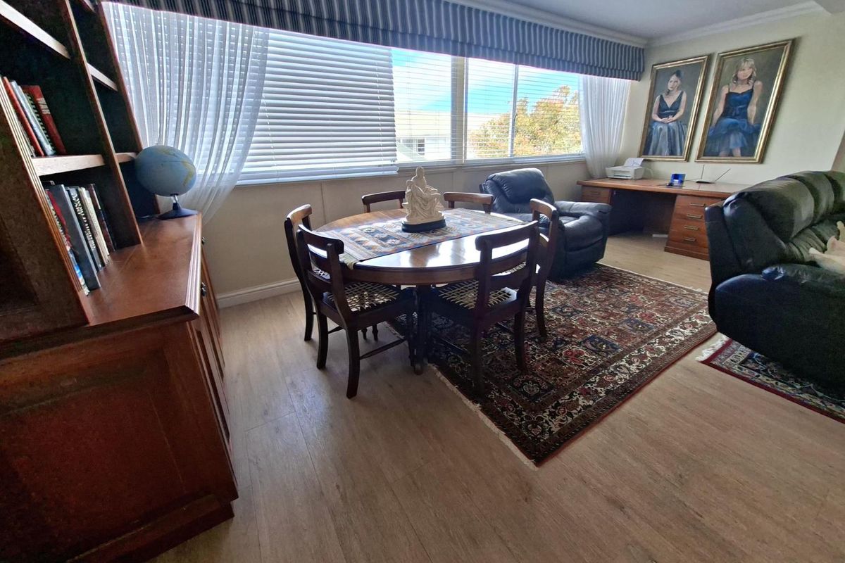A spacious living area featuring light-toned laminate flooring, a built-in desk workspace, and large windows with integrated venetian blinds.