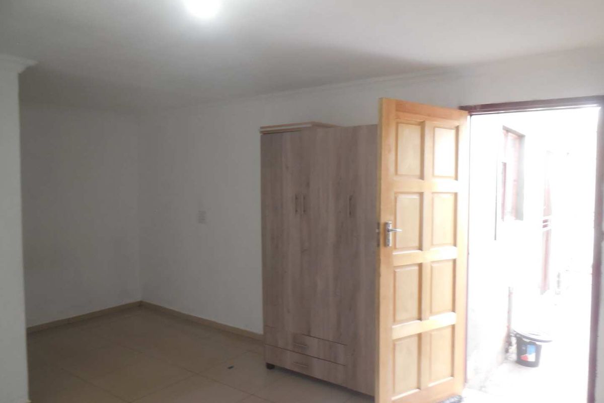 A simple, empty bedroom featuring tiled flooring and a built-in cupboard, with direct access to an outdoor area.