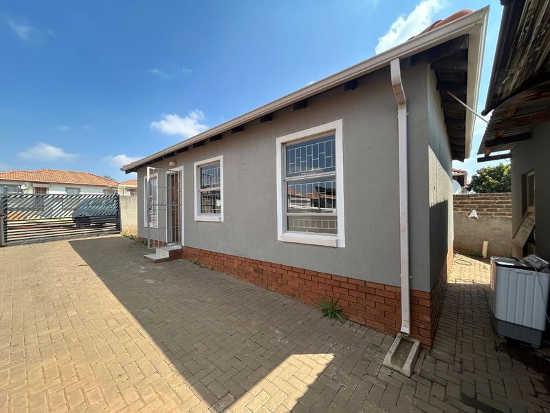 Rear exterior view of the house featuring plastered walls, brick plinth, security bars on windows, and paved outdoor area.