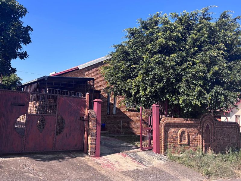 Front exterior view of a face brick house featuring a metal driveway gate, brick boundary wall, and established garden tree.