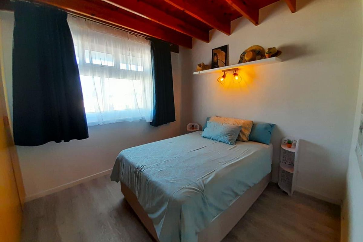 A cozy bedroom featuring exposed timber ceiling beams, light-toned laminate flooring, and a window with curtains.