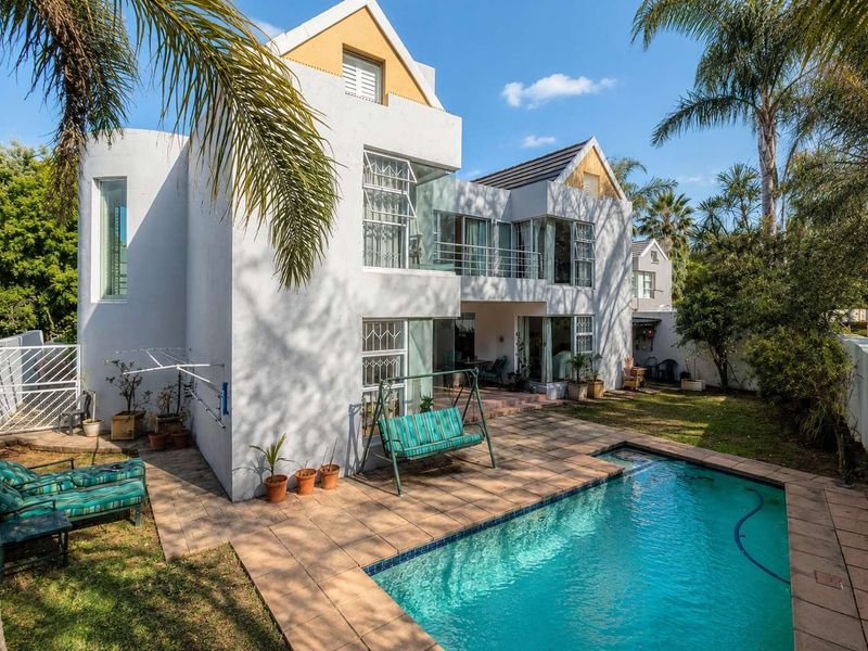 Rear view of a two-story house featuring a swimming pool, tiled patio area, balcony, and established garden surroundings.