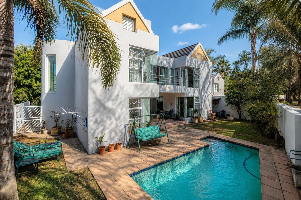 Rear view of a two-story house featuring a swimming pool, tiled patio area, balcony, and established garden surroundings.