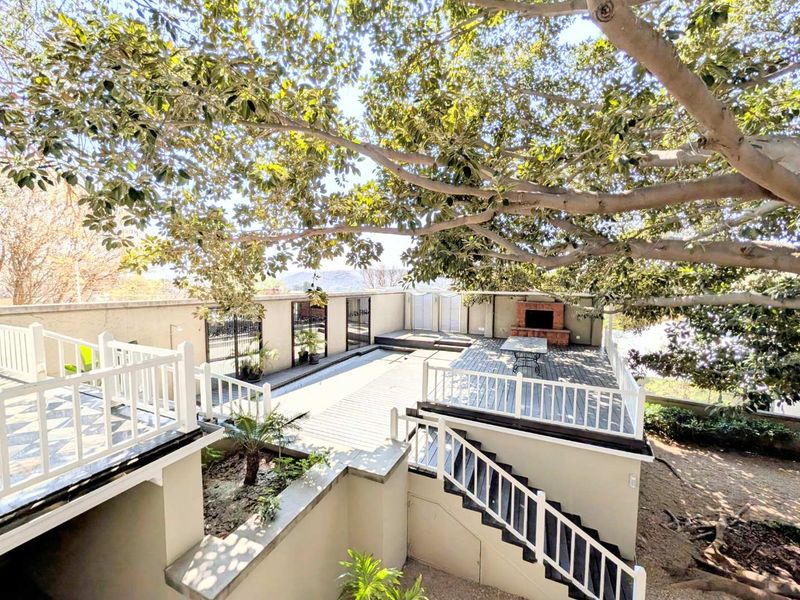 Spacious elevated timber deck patio featuring a built-in brick fireplace, white railings, and stairs, shaded by a large mature tree.