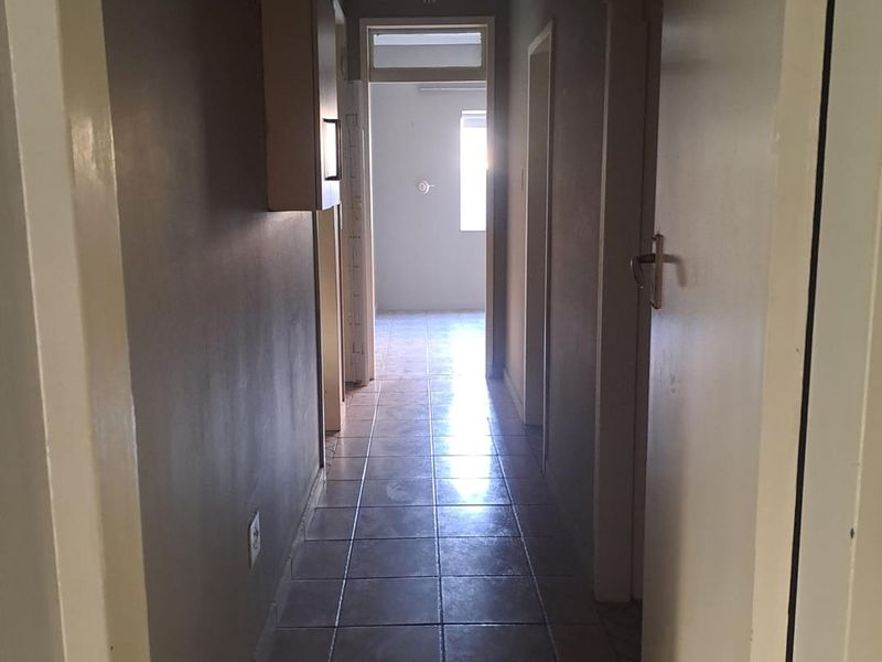 A narrow residential hallway featuring tiled flooring, neutral wall paint, and a central ceiling light fixture, leading towards an adjacent room.
