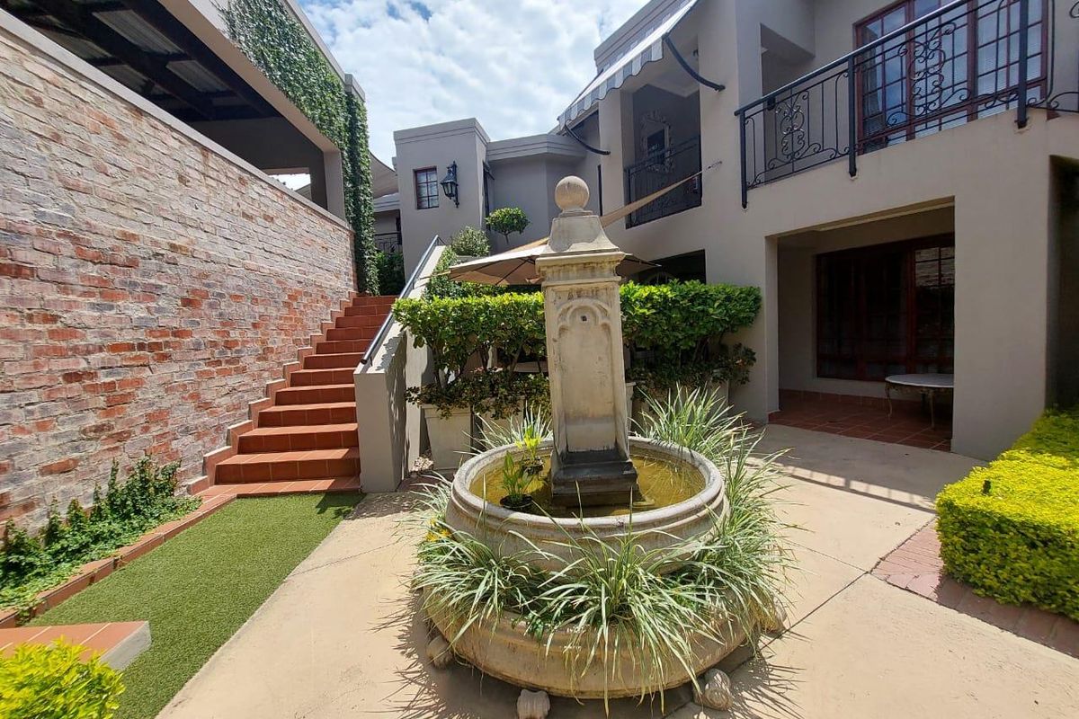 A well-maintained rear courtyard featuring a central stone fountain, brick retaining wall, tiled staircase, and a balcony with wrought iron railings.