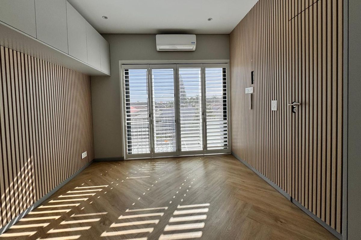 A modern, empty bedroom featuring stylish herringbone timber flooring, feature timber slat wall paneling, built-in overhead storage, and sliding doors with louvre shutters.