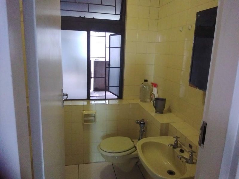 A compact bathroom featuring dated yellow wall tiles, a standard toilet, and a wall-mounted washbasin with a window providing natural light.